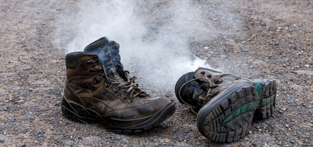 A pair of worn-out work boots on gravel with visible steam or smoke rising from inside, symbolizing extreme heat, sweat, and odor.