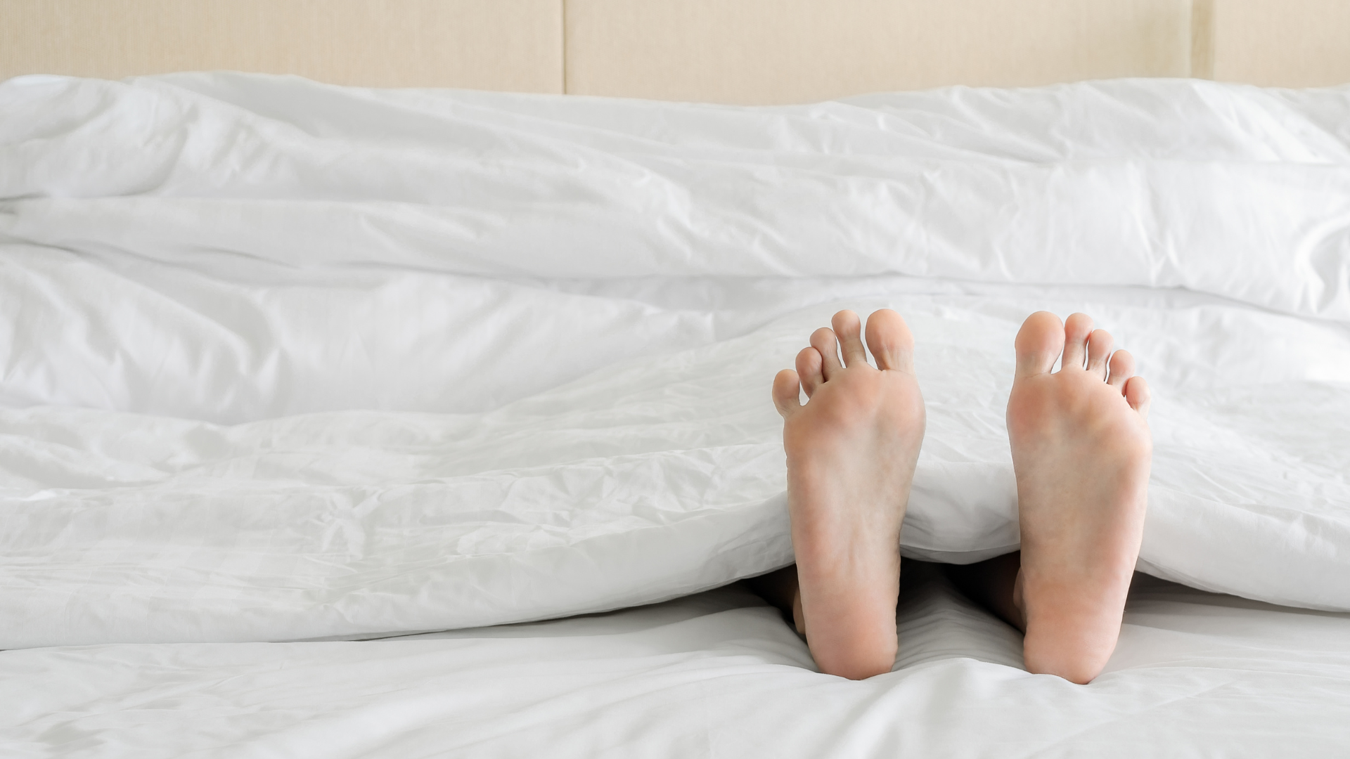 Close-up of a person's bare feet sticking out from under a white duvet on a bed, suggesting relaxation or sleep. The scene conveys comfort, rest, and a focus on foot care or sleep hygiene.