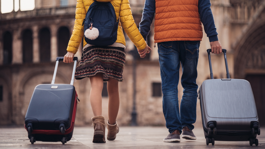 A couple holding hands while walking through a European city square with suitcases. They wear casual outfits with puffer jackets and comfortable shoes, capturing the spirit of travel and exploration.