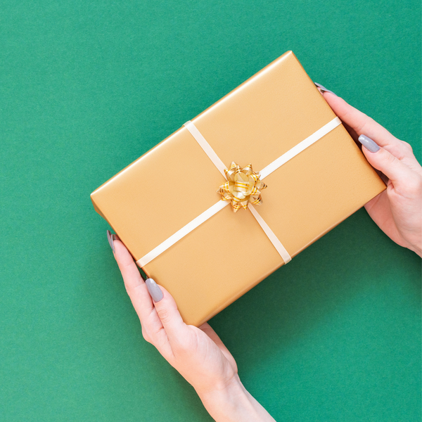 Hands holding a gold-wrapped gift box with a shiny bow on a green background, representing holiday gifting and festive giving season.